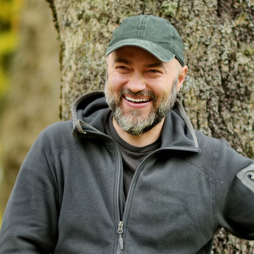 smiling man with beard wearing hat and fleece jacket standing by tree outside