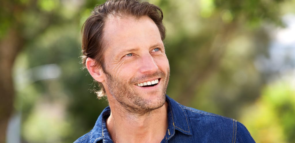 middle aged man wearing denim shirt outside looking up to the right and smiling