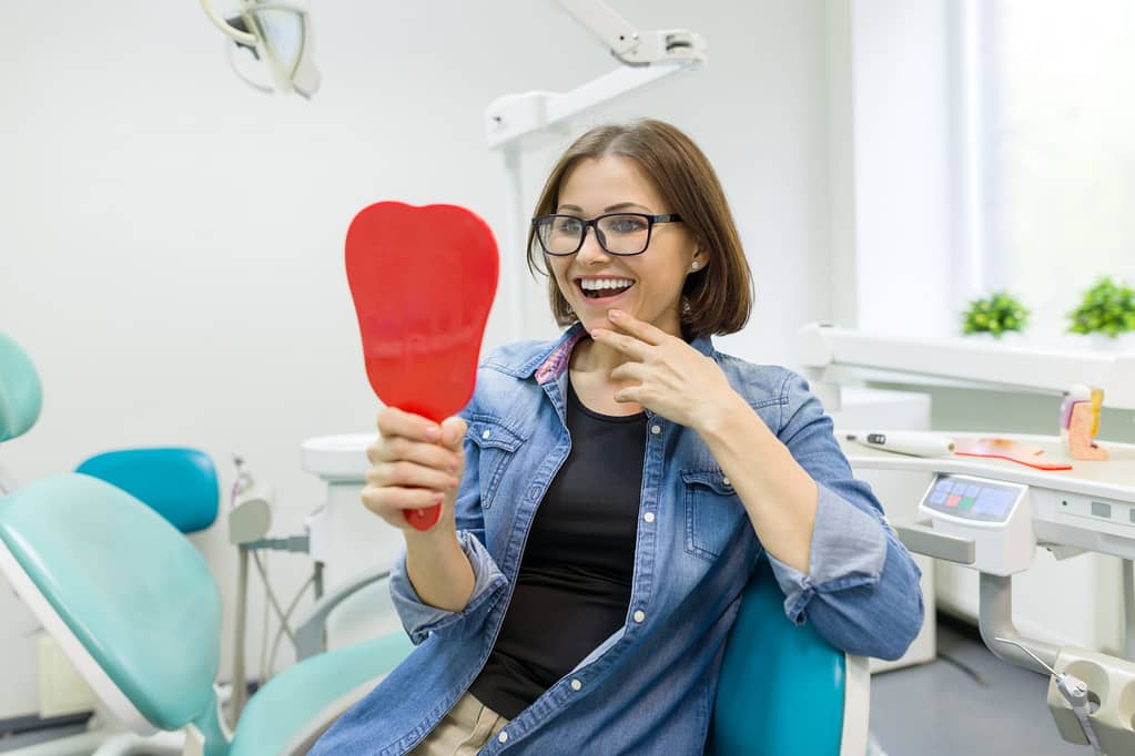 Happy woman patient looking in the mirror at the teeth, sitting in the dental chair.